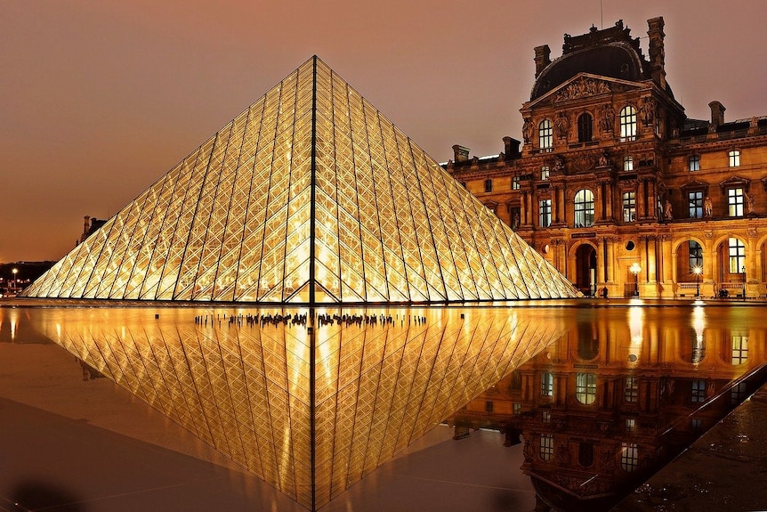 Glass pyramid of the Louvre it up at night, with facade of old building behind.