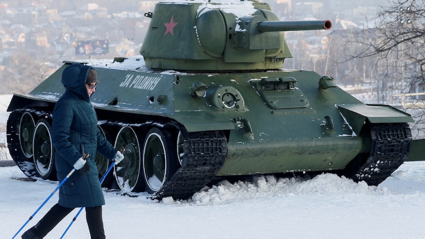 Green army tank with red star sits in deep snow as woman walks past