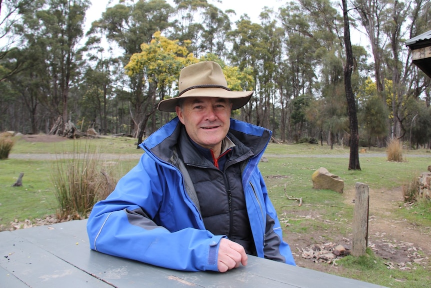 A middle aged man in a hat sits in a bush setting