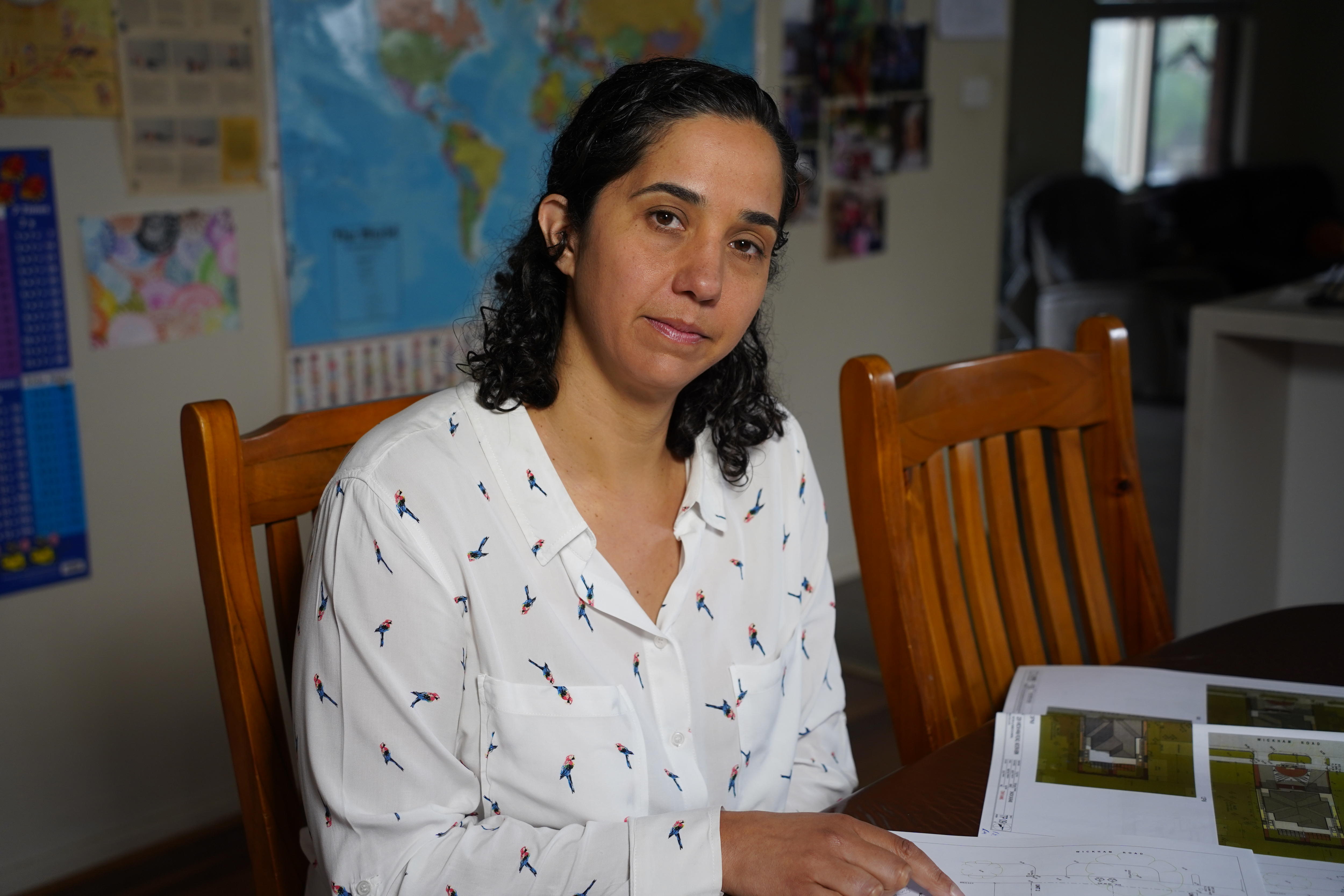 Rebecca sits at a wooden dining table wearing a white shirt, looking at the camera reviewing building plans in front of her.