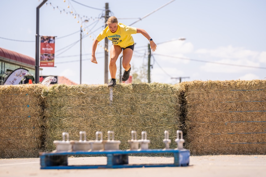 A woman leaps over hay bales piled up on the main street of a country town.