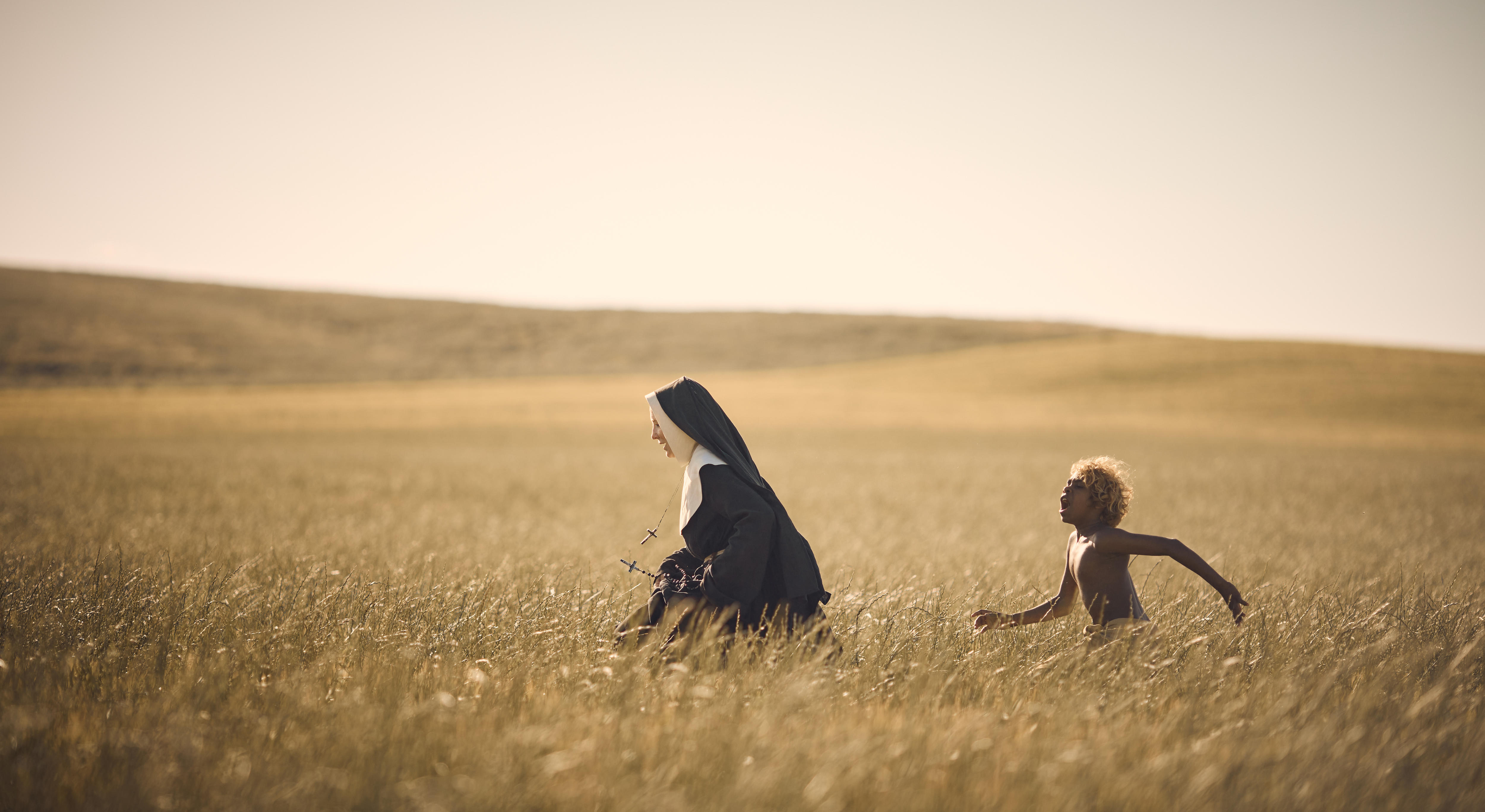 A young Indigenous boy chases a woman in a nun's gown across a wheat field.