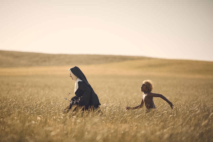 A young Indigenous boy chases a woman in a nun's gown across a wheat field.
