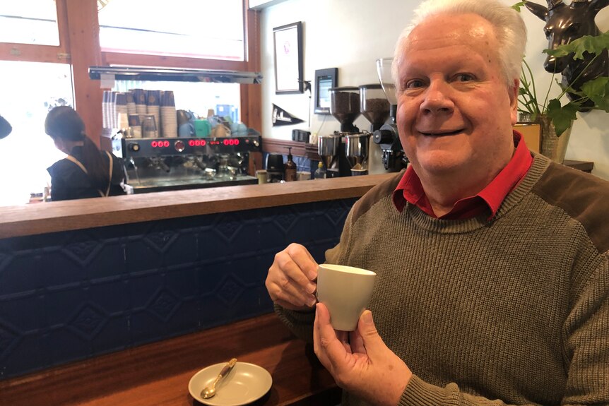 A man sits in a cafe holding a cup of coffee and smiling at the camera.
