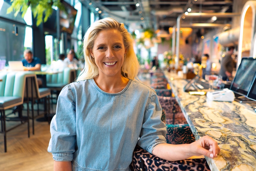 A blonde woman smiles and poses for the camera at a restaurant bar.