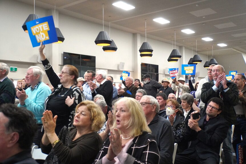 A crowd of No voters clap and hold placards at a Liberal Party forum indoors, some seated and others standing.