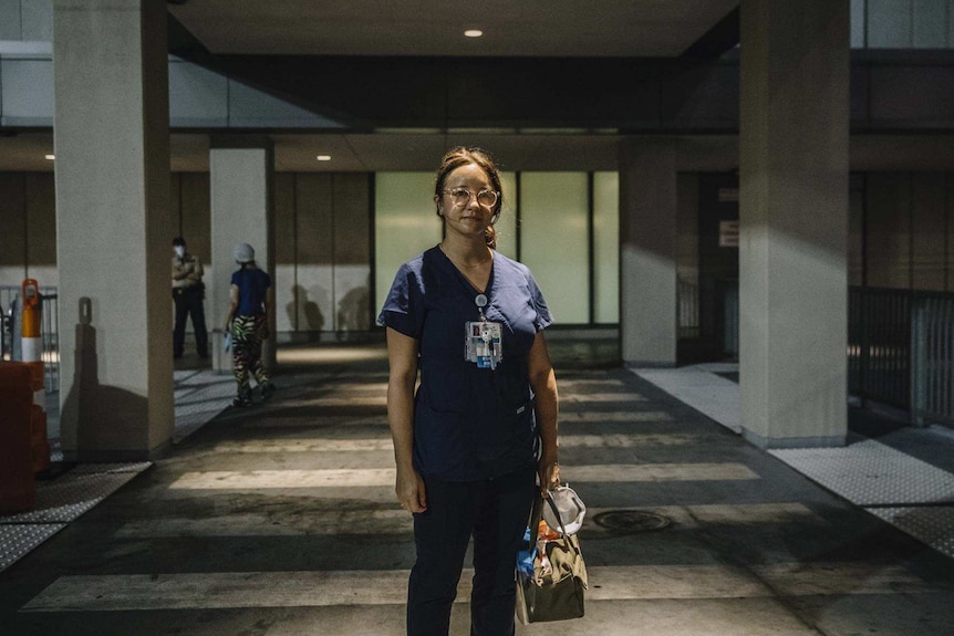 A woman wearing blue scrubs stands outside a medical building.