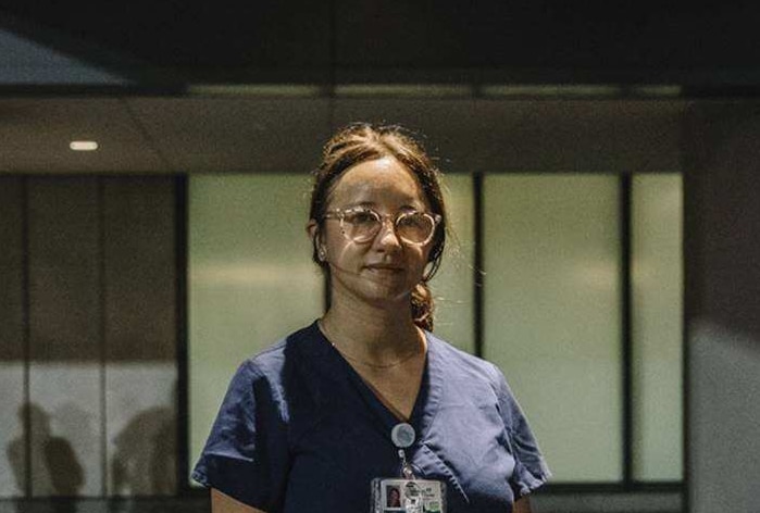 A woman wearing blue scrubs stands outside a medical building.