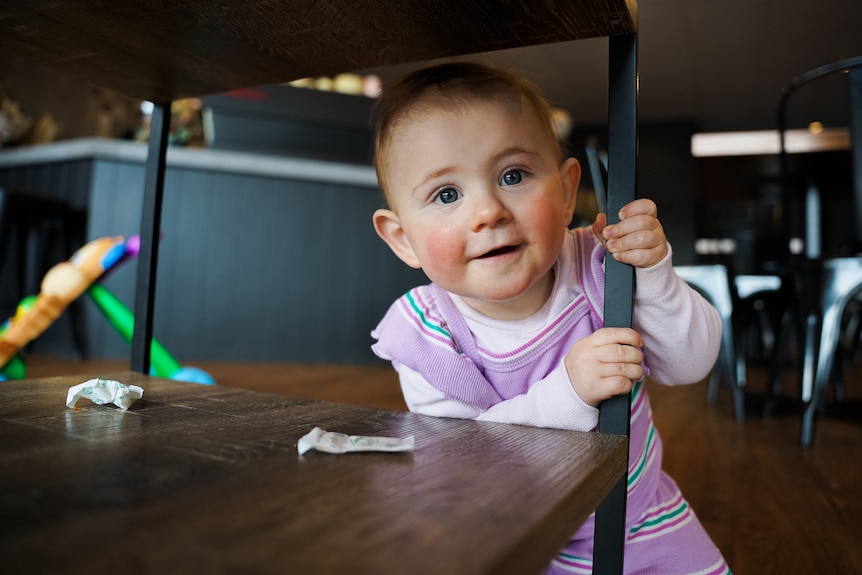 A baby girl in a purple dress smiles from under a table.