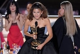 Zendaya holds an Emmy award, an speaks into a microphone. She's wearing a strapless silky glack gown. 