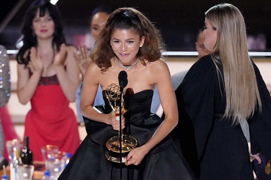 Zendaya holds an Emmy award, an speaks into a microphone. She's wearing a strapless silky glack gown. 