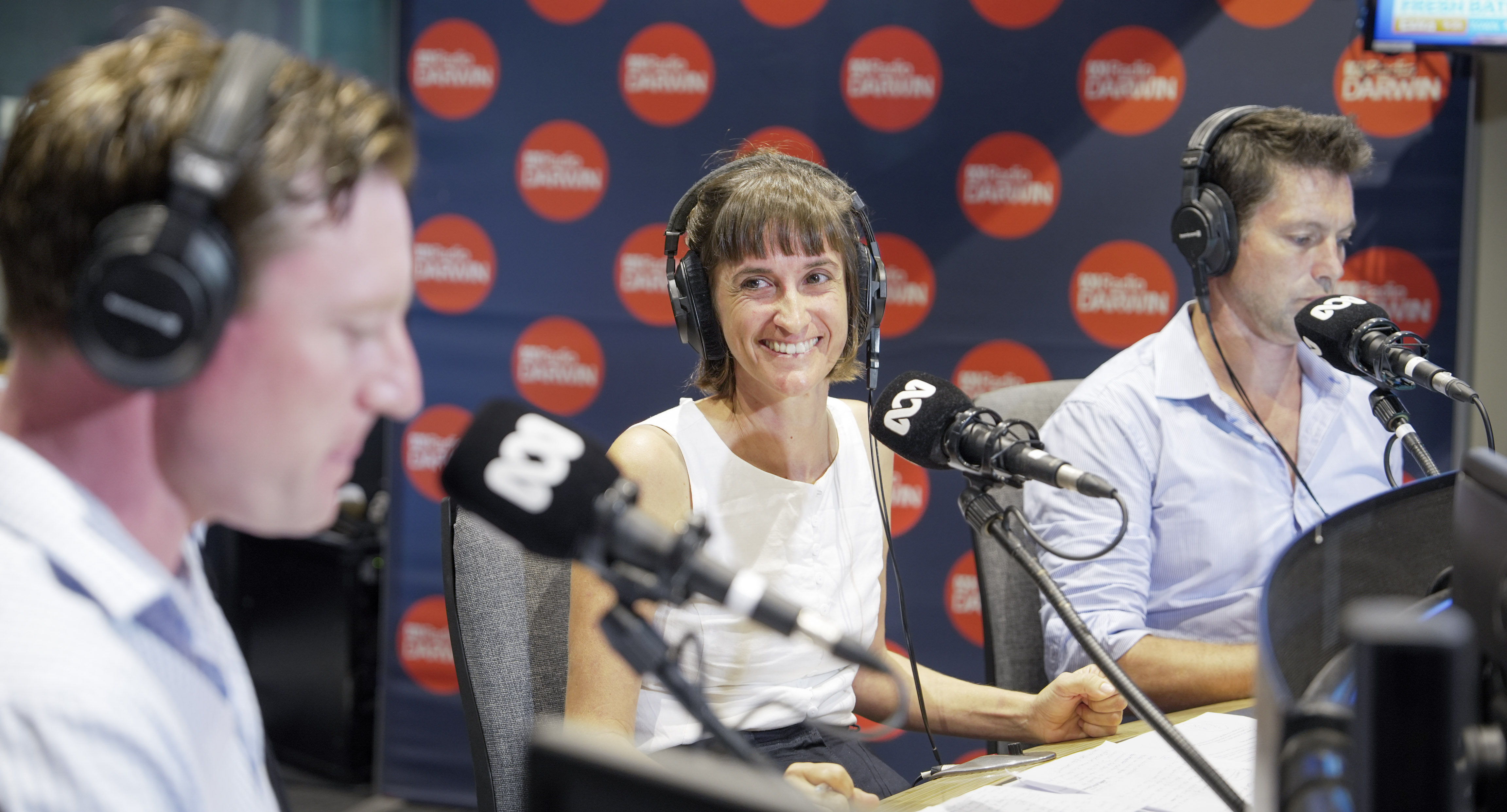 Una mujer sentada frente a un micrófono en un estudio de radio, sonriendo ampliamente, flanqueada por dos hombres.