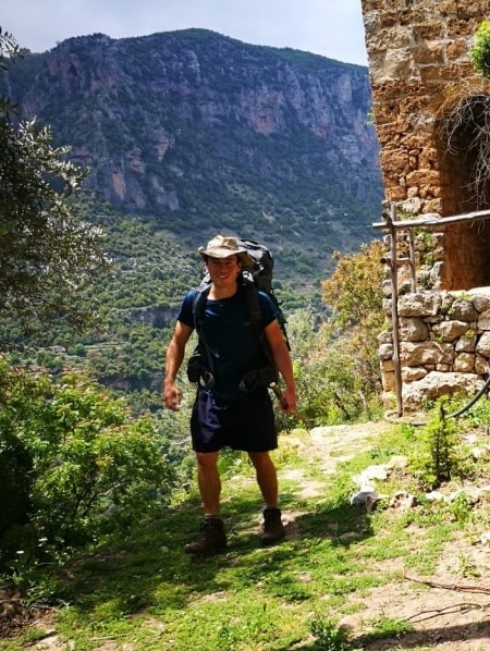 A man in hiking clothing and a hat stands in front of a vast valley in Lebanon.