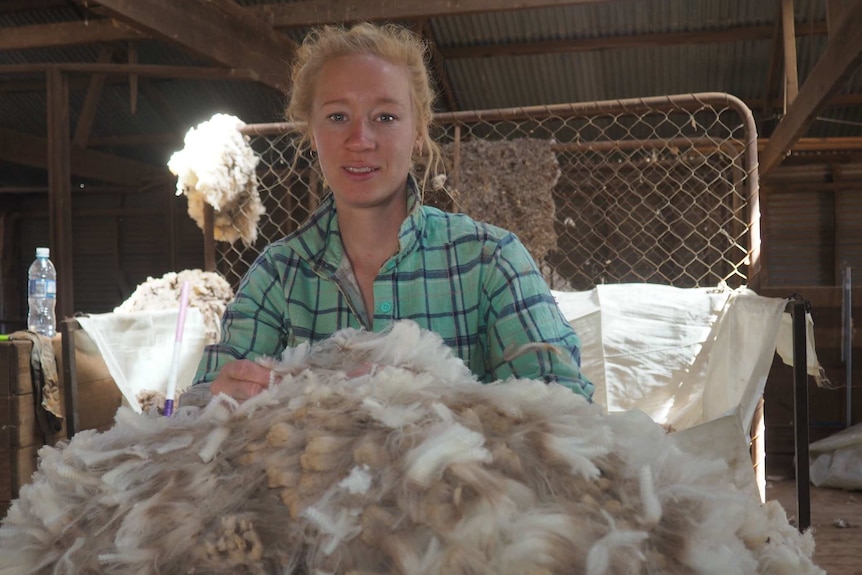 A woman in a flannelette shirt stands behind wool in a shed.