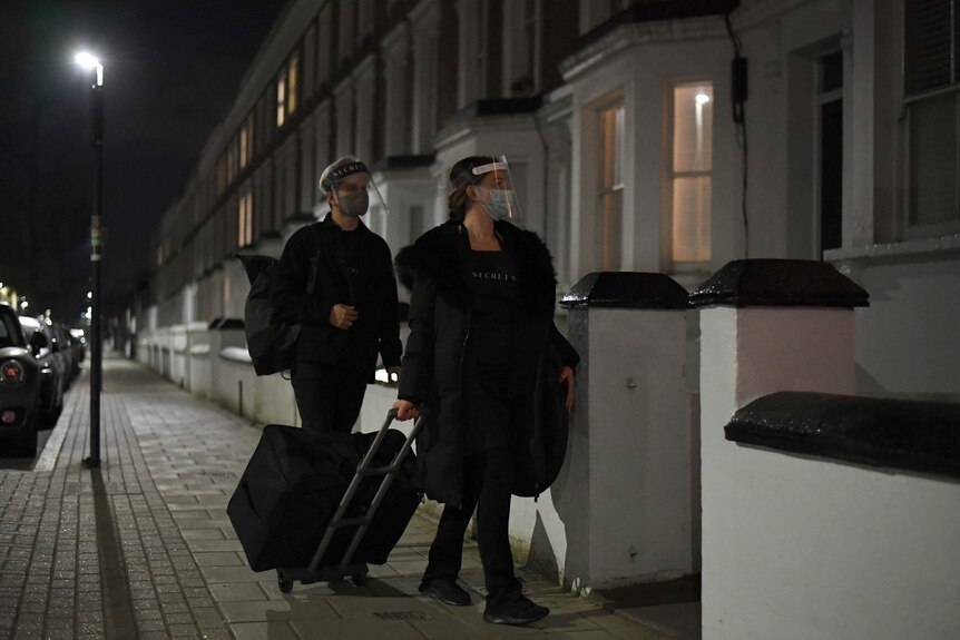 Two people walk on a London street with luggage wearing face shields
