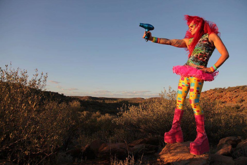A woman with pink hair stands in a field and points a hair dryer away from her. She wears multicoloured tights and top.