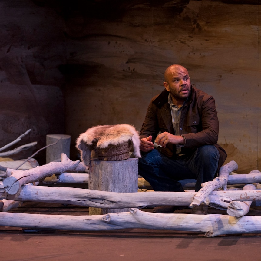 A bald Aboriginal man crouches inside a structure made out of tree branches, next to a tree stump on which a possum skin sits