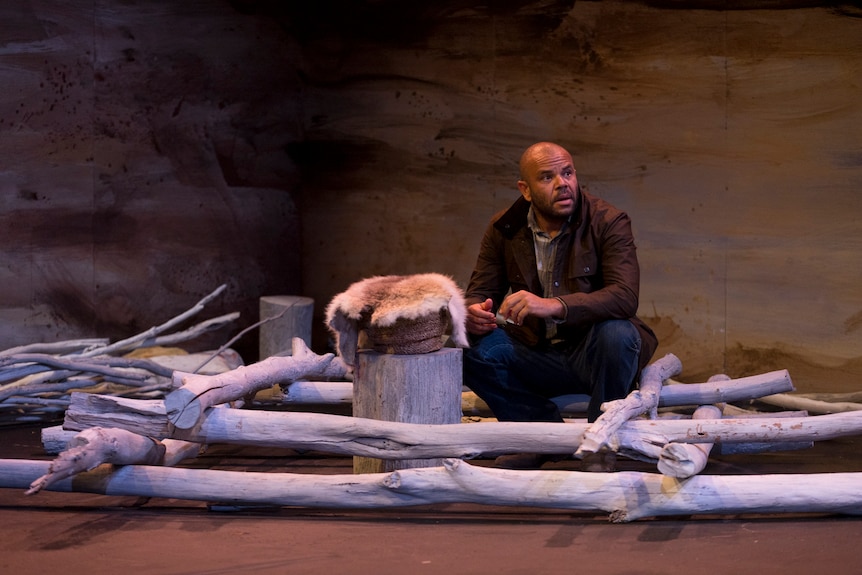 A bald Aboriginal man crouches inside a structure made out of tree branches, next to a tree stump on which a possum skin sits
