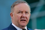 A man with grey hair wearing a navy suit and tie speaking in front of microphones with the background blurred out