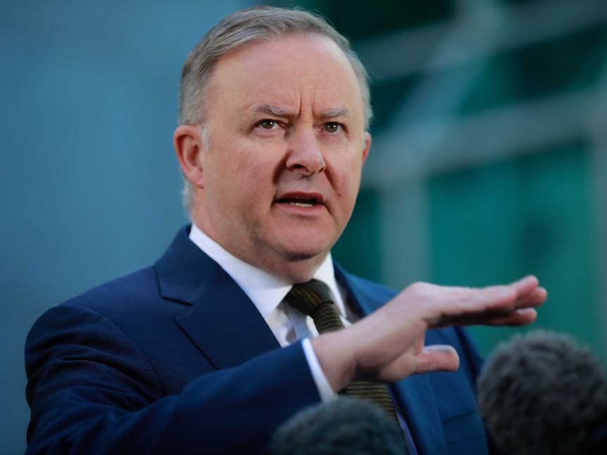 A man with grey hair wearing a navy suit and tie speaking in front of microphones with the background blurred out