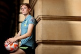 A soccer player wearing a light blue shirt poses with a soccer ball in her hands against a sandstone pillar