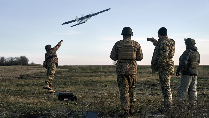 Soldiers stand around as a drone is launched by one of them into the air.