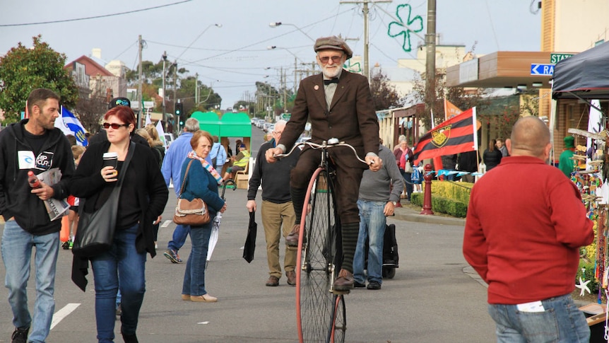 A man riding a penny farthing bike on a street, with other people around him, during a festival.