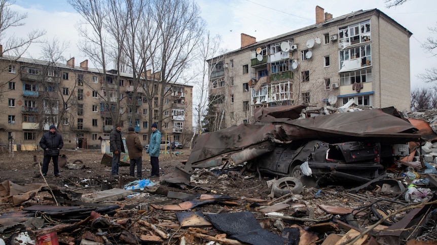 Four local residents stand outside bombed apartment buildings with debris piled on street.