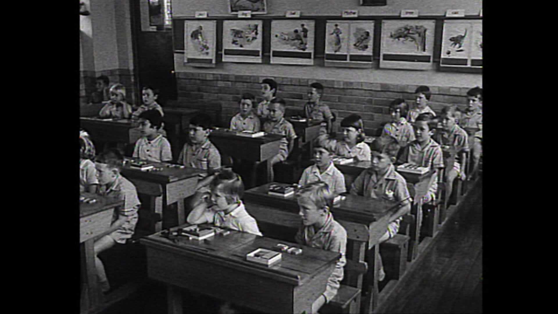 A black and white image of a classroom full of young students sitting at old wooden desks in the 1960s