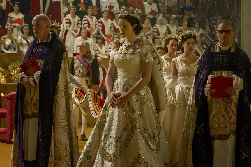 Claire Foy, as the Queen, wears a crown and elaborate white dress at her coronation ceremony.