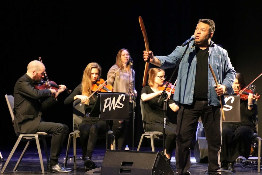 Phil Walleystack holds a stick and boomerang on stage, with orchestra members in the background.