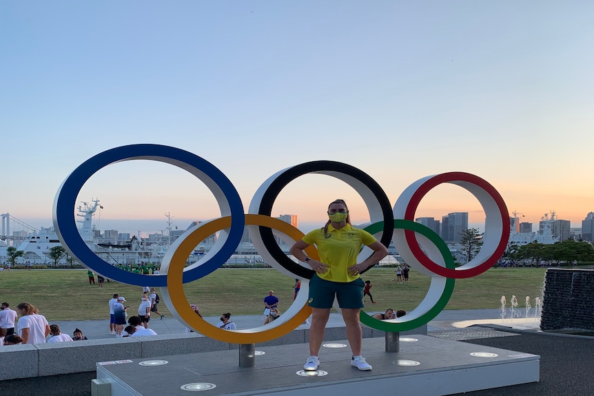 A woman in an Australian Olympic uniform standing in front of the Olympic rings statue.