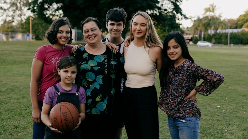 Five young people and a woman stand in a park embracing eachother