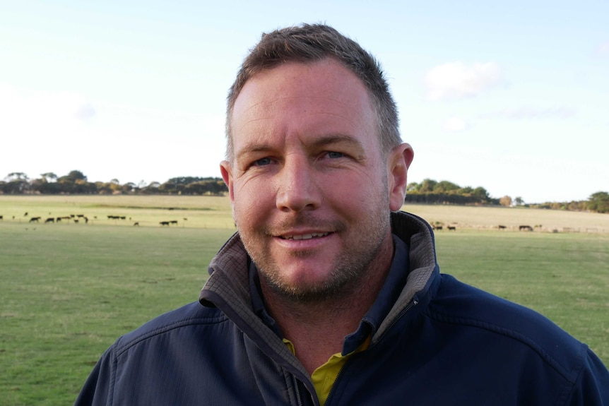 A head and shoulders image of a man standing on a farm with livestock seen grazing in the background.