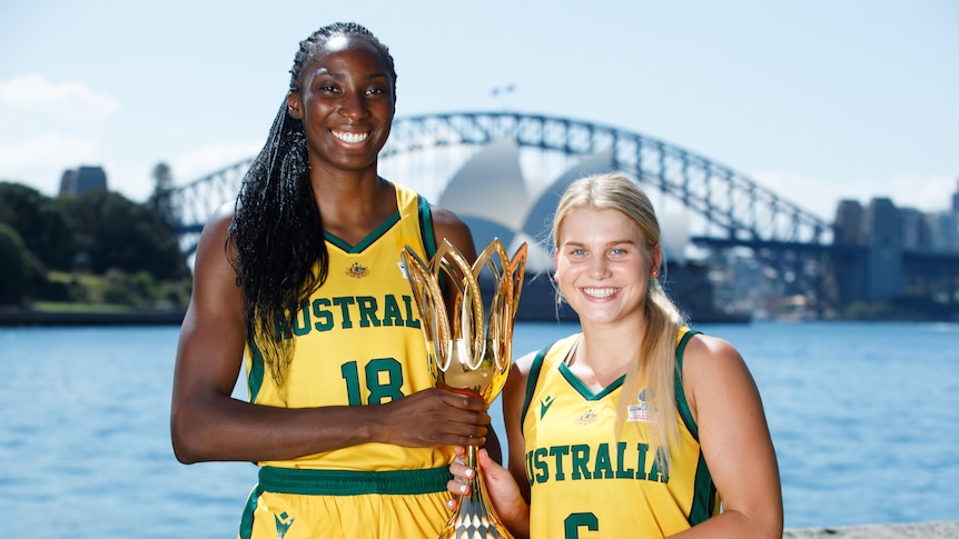 Two Australian women's basketballers hold the World Cup trophy in front of Sydney Harbour, smiling