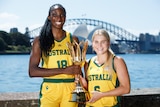 Two Australian women's basketballers hold the World Cup trophy in front of Sydney Harbour, smiling
