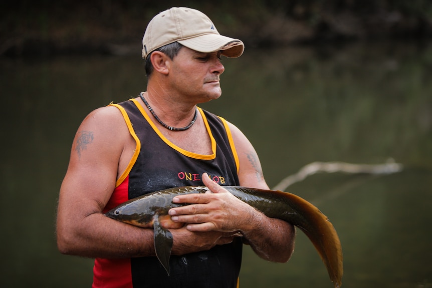 Indigenous man in a black singlet and a beige cap holding a long fish in his arms, looking wistful.