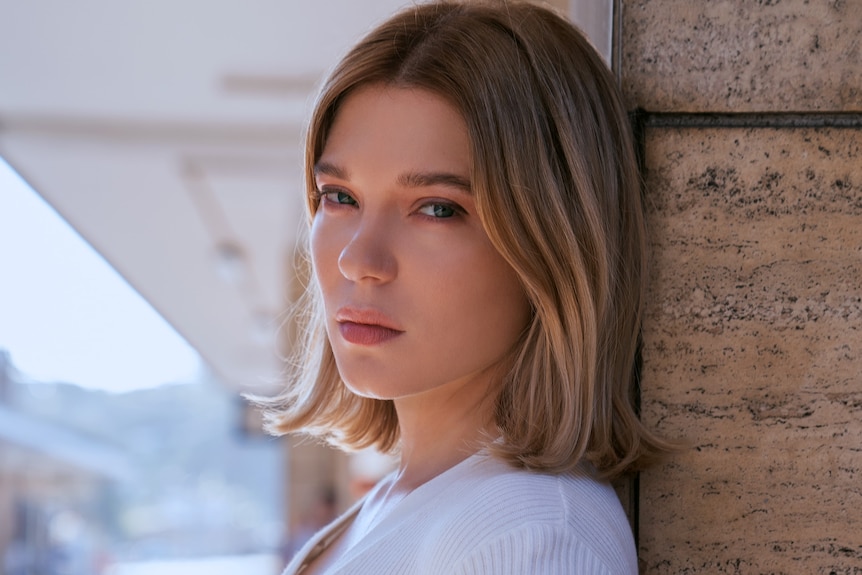 A blonde woman in her 30s turns her head to the side to look straight into the camera. She is leaning against a sandstone wall.