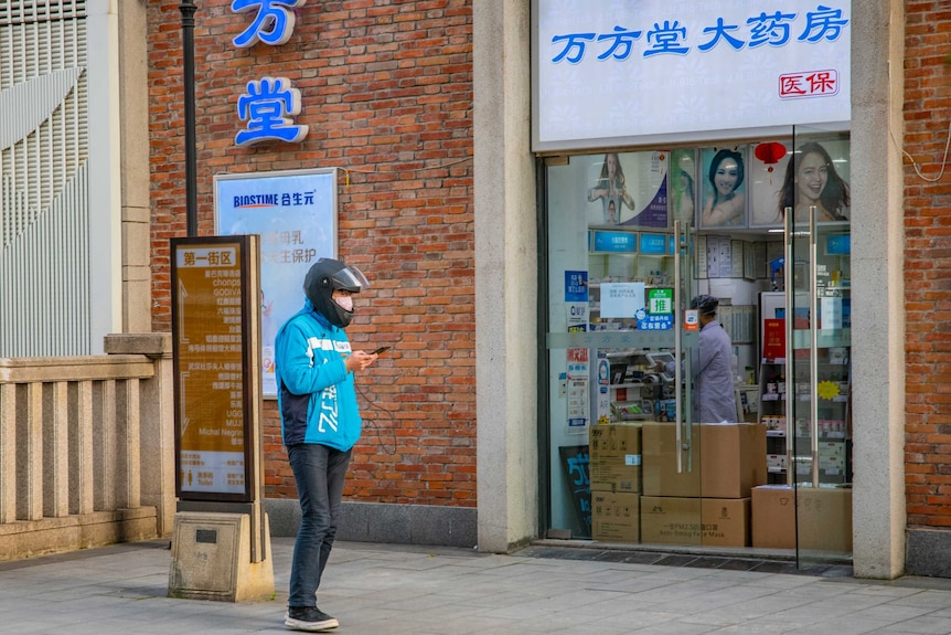 A man in a blue jacket looks at his phone in front of a pharmacy where lots of cardboard boxes are stacked.