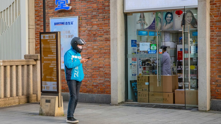 A man in a blue jacket looks at his phone in front of a pharmacy where lots of cardboard boxes are stacked.