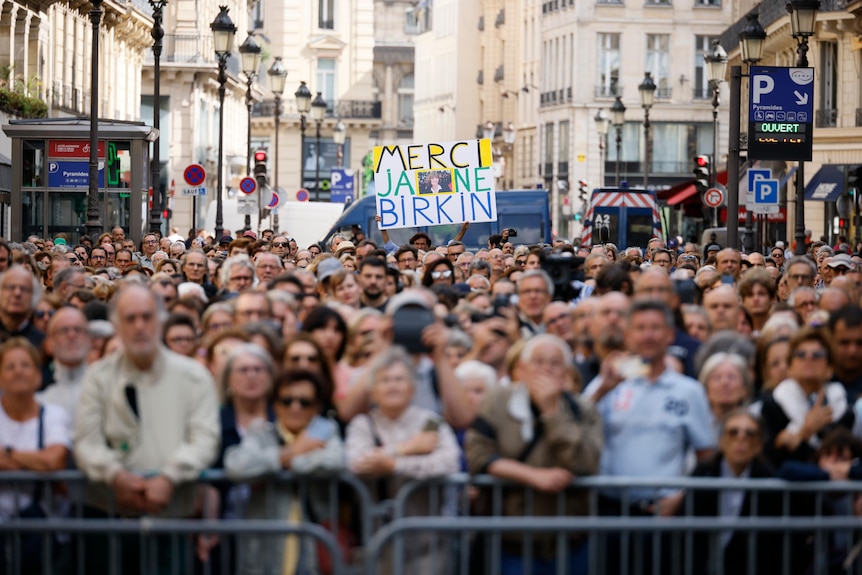 a large crowd gather in front of a barrier with a sign that reads thankyou jane birkin in French 