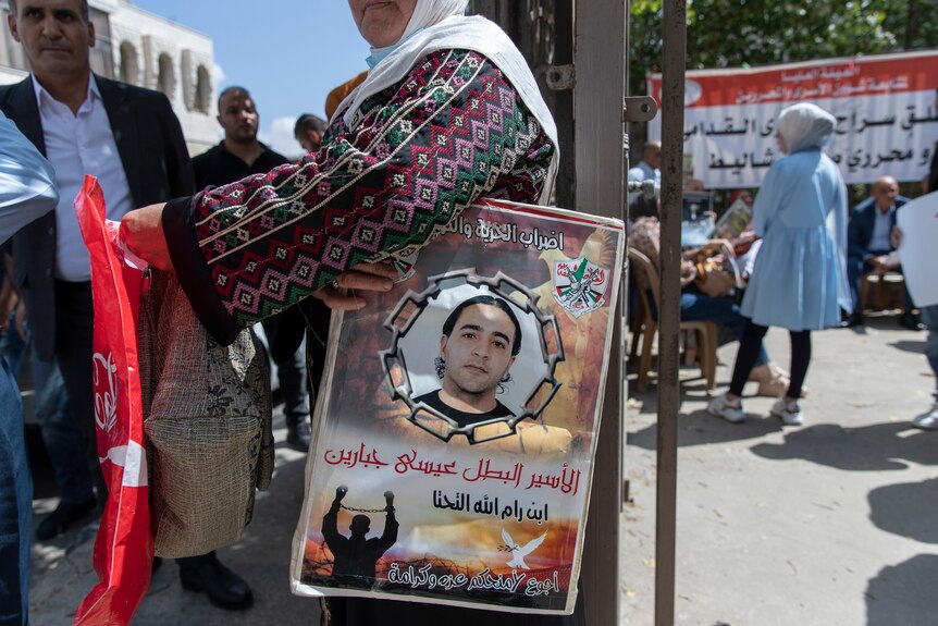 A woman wearing a head scarf holds a poster showing a young Palestinian man surrounded by Arabic writing.