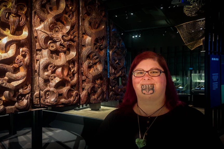 A woman with ta moko, traditional maori facial tattoo, stands next to a display of large wooden carvings. 