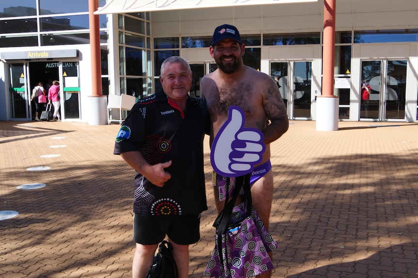 A man wearing purple swim briefs poses with a traveller at airport.