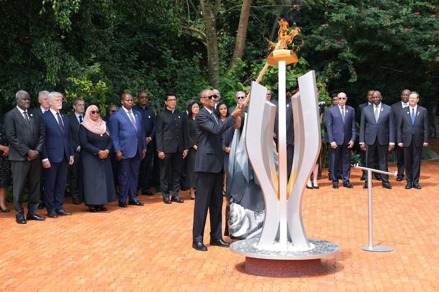 A thin, older African man in a suit lights a flame atop a silver sculpture with a long torch, as others in suits look on.