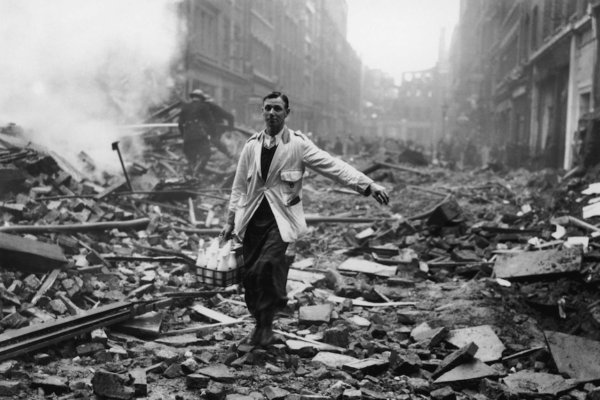 A historic photo of a milkman walking over rumble to deliver milk as firemen put out fires in the background.