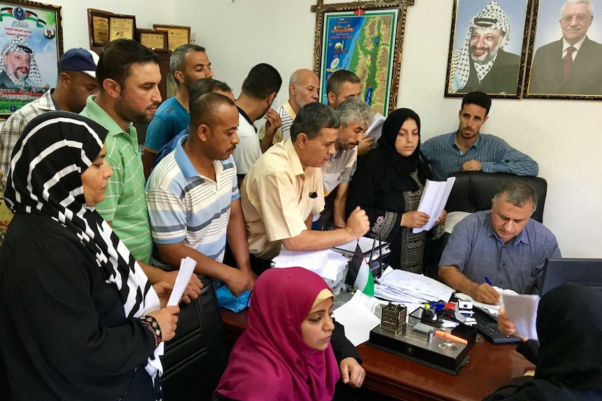 A crowd of people, many clutching papers, gather around a man sitting at a desk filing in forms.