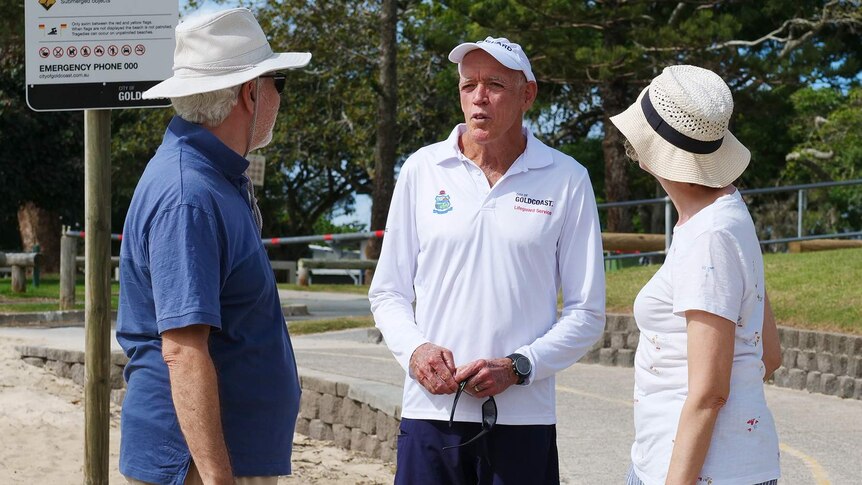 Older man white lifeguard shirt on and white cap on talks to mand and woman both wearing hats with sand and a path in background