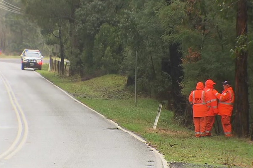 Three workers in bright orange SES uniforms stand near a wet road, with a police car in the background.