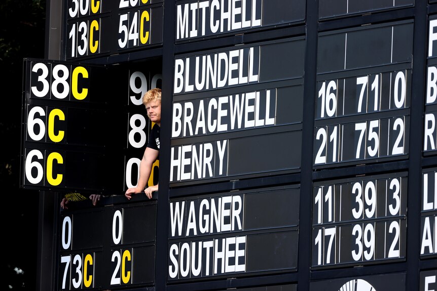 A man looks out a window panel in a cricket scoreboard during a Test match, as the bowling figures can be seen next to him.